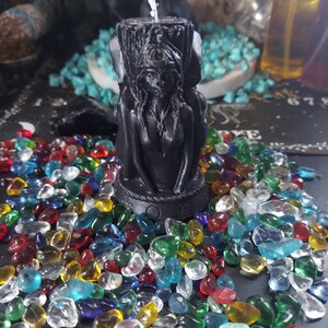 May include: Black candle with a female figure sculpted into the wax. The candle is surrounded by colorful glass pebbles.