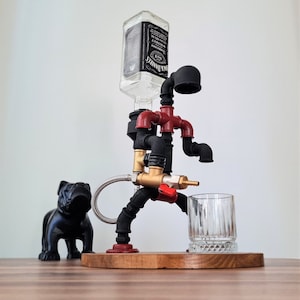 May include: A black metal liquor dispenser shaped like a walking figure with red and gold accents. The dispenser has a clear glass bottle on top and a glass shot glass in front. A black bulldog figurine is on the left side of the dispenser. The dispenser is on a wooden base.