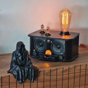 May include: A black, hooded figure figurine sits next to a black, rectangular electronic device featuring two circular speakers, a central analog gauge with an orange glow, two small vacuum tubes, and a large, glowing Edison-style light bulb.