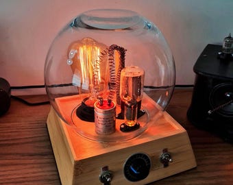 May include: A table lamp featuring a clear glass dome housing an Edison bulb, a cylindrical capacitor labeled AI-Elke, and a vacuum tube. The base is made of light wood and includes two metal toggle switches and a central black rotary control knob.
