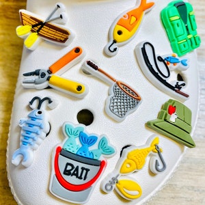 May include: A set of 11 colorful fishing-themed shoe charms. The charms include a fishing rod, a fishing net, a bucket of bait, a life vest, a lure, a fish, and a canoe. The charms are made of rubber and are designed to be attached to shoes.