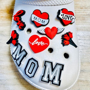 May include: White Crocs shoe with red and black heart-shaped charms. The charms have the words "love", "MOM", and "LOVE" on them. There are also two charms with wings and a charm with a flower.