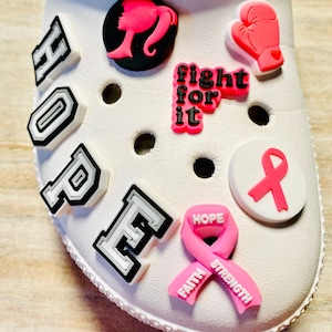 May include: White Crocs shoe decorated with various charms. Charms include the word "HOPE", a pink ribbon, a silhouette, a boxing glove, and the phrase "fight for it". The charms are in shades of pink, black, and white.