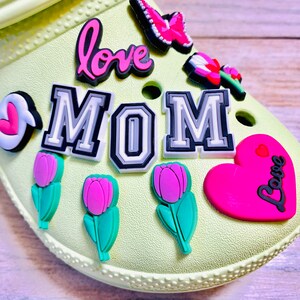 May include: A light green shoe with various colorful charms attached. The charms include a pink heart with the word "Love" in black, a pink flower, a pink butterfly, a black and white charm that says "MOM", a pink flower with a green stem, and a pink heart with the word "Love" in black.