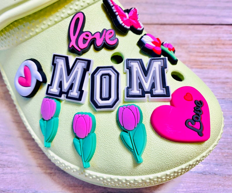 May include: A light green shoe with various colorful charms attached. The charms include a pink heart with the word "Love" in black, a pink flower, a pink butterfly, a black and white charm that says "MOM", a pink flower with a green stem, and a pink heart with the word "Love" in black.
