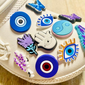 Pode incluir: Um sapato branco com v&aacute;rios pingentes presos a ele. Os pingentes incluem um olho turco azul, uma m&atilde;o branca com um olho turco, uma flor de lavanda, uma borboleta azul, um s&iacute;mbolo yin yang azul e branco, uma pomba branca, uma pena azul e um olho azul com c&iacute;lios laranja.