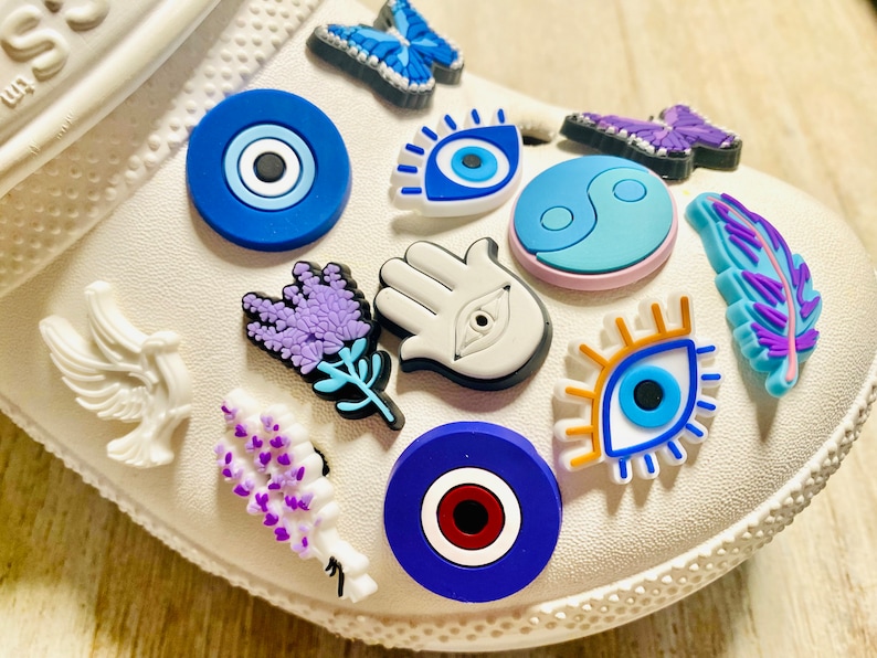 Pode incluir: Um sapato branco com v&aacute;rios pingentes presos a ele. Os pingentes incluem um olho turco azul, uma m&atilde;o branca com um olho turco, uma flor de lavanda, uma borboleta azul, um s&iacute;mbolo yin yang azul e branco, uma pomba branca, uma pena azul e um olho azul com c&iacute;lios laranja.