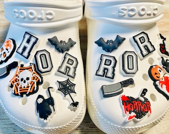 May include: White clogs with Halloween-themed shoe charms. The charms include a spider web with a skull, a ghost, a bat, a knife, a pumpkin, a heart with a knife, and the letters "HORROR" in black and gray.