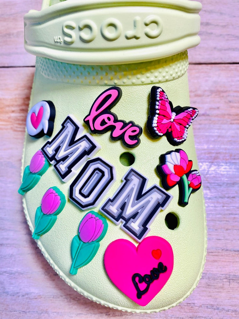 May include: A pair of green Crocs with various colorful charms attached. The charms include a heart with the word "love", a butterfly, a flower, a tulip, and a large charm that says "MOM" in black and white letters.