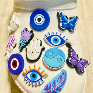 Pode incluir: Um sapato branco com v&aacute;rios amuletos coloridos presos. Os amuletos incluem um olho turco azul, uma borboleta roxa, uma m&atilde;o branca com um olho, um s&iacute;mbolo yin yang azul e branco, um olho turco vermelho e azul, um olho azul com c&iacute;lios, uma flor roxa, uma pena roxa e uma borboleta azul.