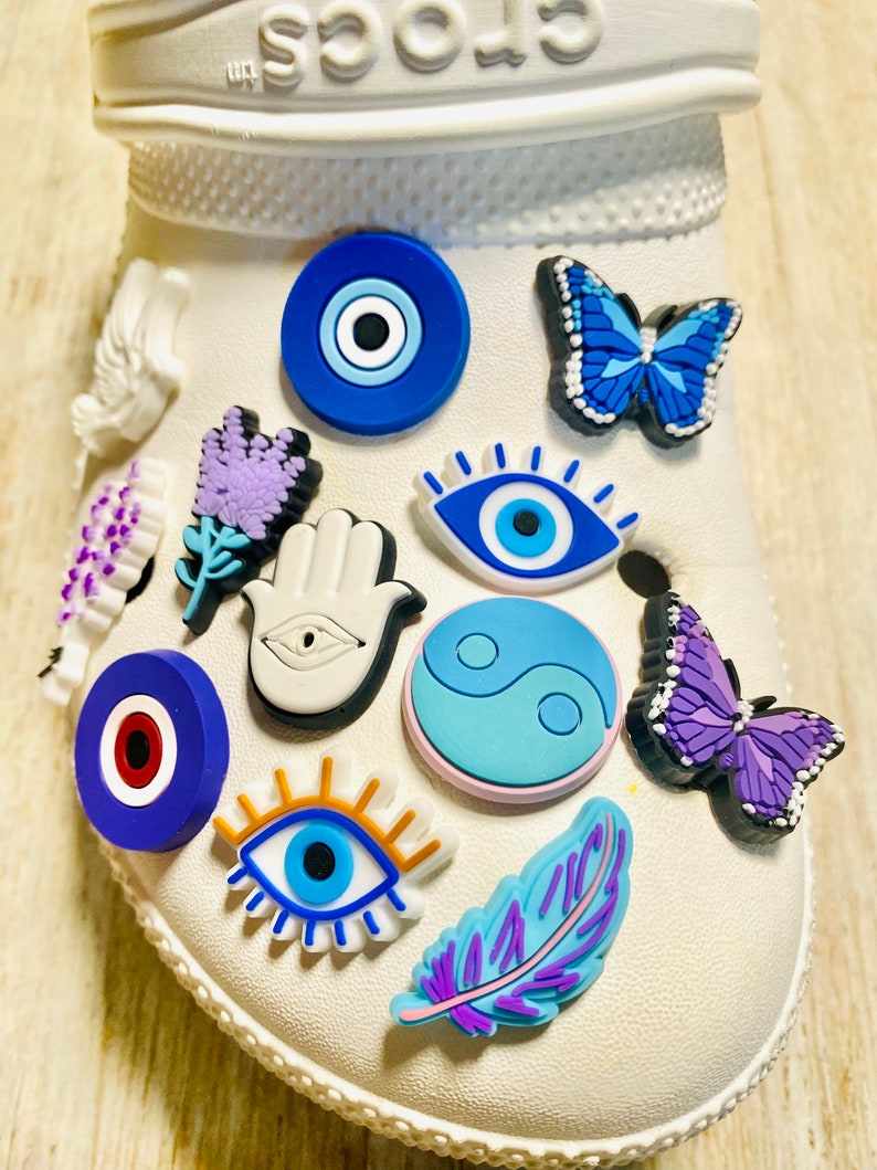 Pode incluir: Um sapato branco com v&aacute;rios amuletos coloridos presos. Os amuletos incluem um olho turco azul, uma borboleta roxa, uma m&atilde;o branca com um olho, um s&iacute;mbolo yin yang azul e branco, um olho turco vermelho e azul, um olho azul com c&iacute;lios, uma flor roxa, uma pena roxa e uma borboleta azul.
