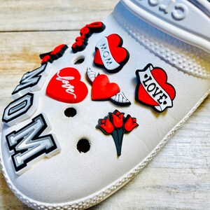 May include: White clog shoe with various colorful charms, including red hearts with the words "Love", "Mom", and "Love" in a tattoo style, a red heart with wings, and a red heart with the word "Love" in cursive. The shoe also has a charm that says "Mom" in white letters on a black background.