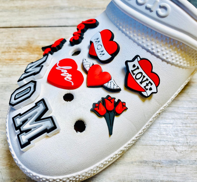 May include: White clog shoe with various colorful charms, including red hearts with the words "Love", "Mom", and "Love" in a tattoo style, a red heart with wings, and a red heart with the word "Love" in cursive. The shoe also has a charm that says "Mom" in white letters on a black background.