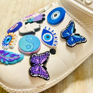 Pode incluir: Sapato branco com v&aacute;rios amuletos coloridos, incluindo olhos turcos azuis e brancos, uma borboleta roxa, uma pena roxa, um s&iacute;mbolo yin yang e um amuleto de m&atilde;o roxo.