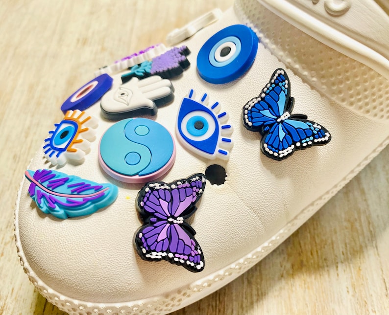 Pode incluir: Sapato branco com v&aacute;rios amuletos coloridos, incluindo olhos turcos azuis e brancos, uma borboleta roxa, uma pena roxa, um s&iacute;mbolo yin yang e um amuleto de m&atilde;o roxo.