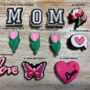 May include: A set of pink and white shoe charms with various designs, including letters spelling "MOM", flowers, hearts, a butterfly, and the word "love".