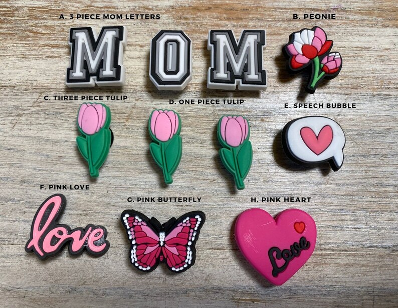 May include: A set of pink and white shoe charms with various designs, including letters spelling "MOM", flowers, hearts, a butterfly, and the word "love".