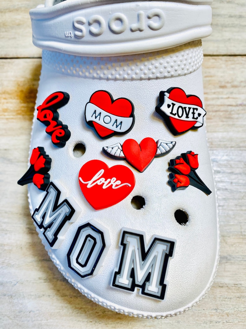 May include: White Crocs shoe with red and black heart-shaped charms. The charms have the words "love", "MOM", and "LOVE" on them. There are also two charms with wings and a charm with a flower.