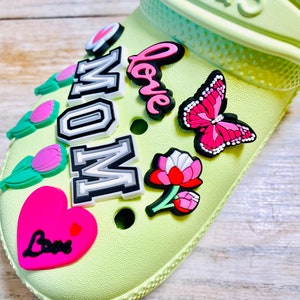 May include: A light green shoe with various colorful charms attached. The charms include a pink heart with the word "Love" in black, a pink butterfly, a pink flower, a green tulip, and a white and gray charm that says "MOM" in black letters.
