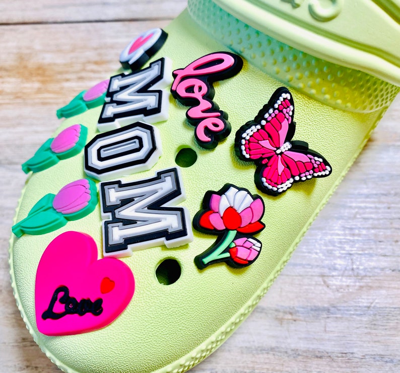 May include: A light green shoe with various colorful charms attached. The charms include a pink heart with the word "Love" in black, a pink butterfly, a pink flower, a green tulip, and a white and gray charm that says "MOM" in black letters.