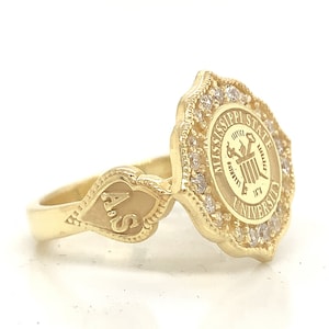 May include: A gold-colored ring featuring the Mississippi State University seal, surrounded by small, clear stones. The ring has a decorative band with the initials "A.S." engraved on it. The ring is set against a white background.