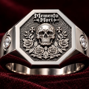 May include: A silver-toned ring featuring an octagonal face with a central embossed skull, roses, and two small crosses. The text Memento Mori is engraved at the top. A clear, round, faceted stone is set into each side of the polished metal band.