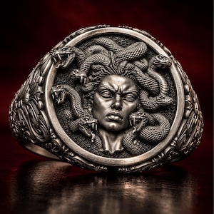 May include: A silver ring featuring a detailed relief of a face surrounded by multiple snakes. The circular centrepiece displays a central figure with closed eyes and textured scales, while the band is decorated with intricate scrollwork patterns.