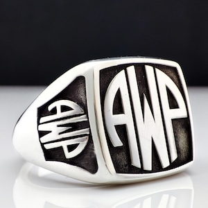 May include: A silver ring with a rectangular face featuring a monogram of the letters "AWP" in a circle. The letters are raised and white against a black background.