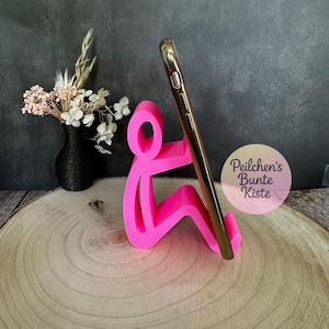 Pu&ograve; includere: Un supporto per telefono rosa acceso a forma di persona che tiene in mano un telefono dorato. Il supporto &egrave; su una superficie di legno, con un vaso nero di fiori secchi sullo sfondo. Il testo "Peilchen's Bunte Kiste" &egrave; visibile.