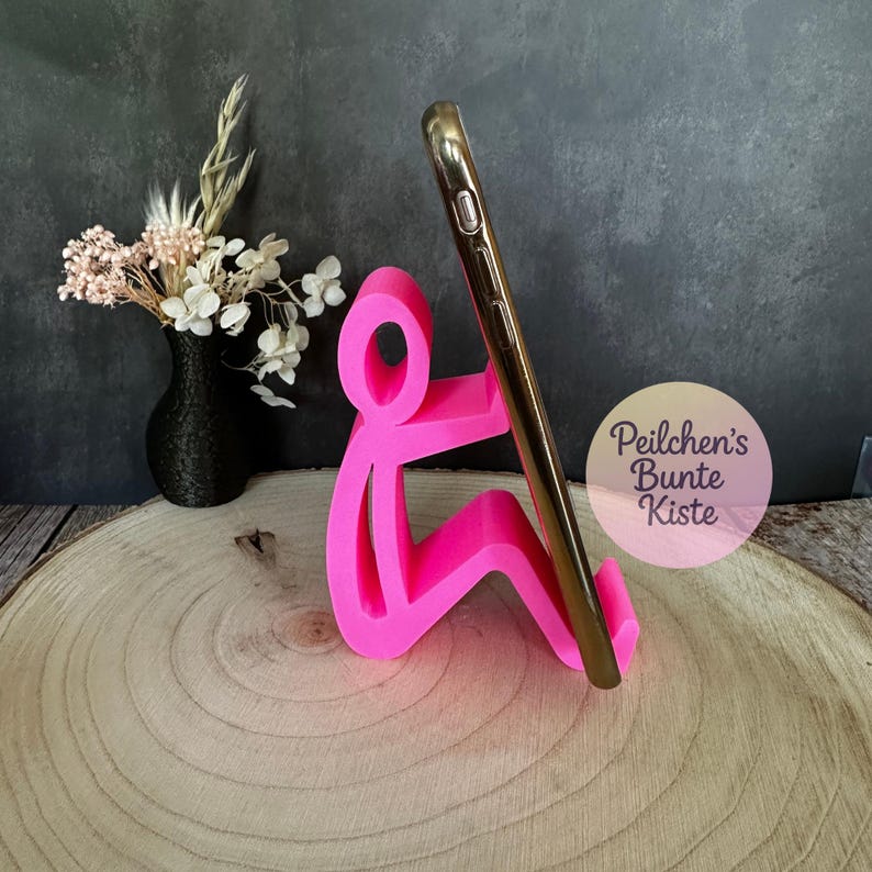 Pu&ograve; includere: Un supporto per telefono rosa acceso a forma di persona che tiene in mano un telefono dorato. Il supporto &egrave; su una superficie di legno, con un vaso nero di fiori secchi sullo sfondo. Il testo "Peilchen's Bunte Kiste" &egrave; visibile.