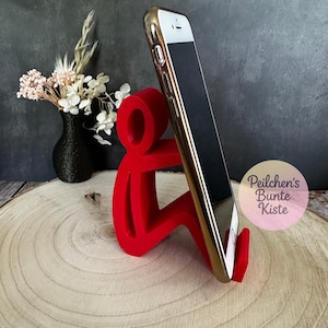 Pu&ograve; includere: Un supporto per telefono rosso a forma di persona che tiene in mano uno smartphone con cornice dorata. Il telefono ha uno schermo nero ed &egrave; angolato. Il supporto &egrave; su una superficie di legno, con un vaso nero e fiori sullo sfondo.