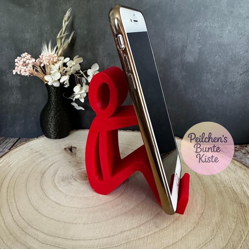Pu&ograve; includere: Un supporto per telefono rosso a forma di persona che tiene in mano uno smartphone con cornice dorata. Il telefono ha uno schermo nero ed &egrave; angolato. Il supporto &egrave; su una superficie di legno, con un vaso nero e fiori sullo sfondo.