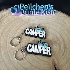 May include: Two black and white keyrings with silver key rings. Each keyring has the words "happy camper" written in white with a teal blue outline.