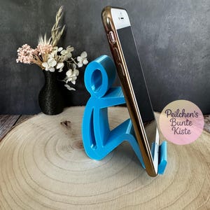 Pu&ograve; includere: Un supporto per telefono blu brillante a forma di persona che tiene in mano un telefono dorato e nero. Il supporto &egrave; su una superficie di legno. Un vaso nero con fiori &egrave; sullo sfondo.