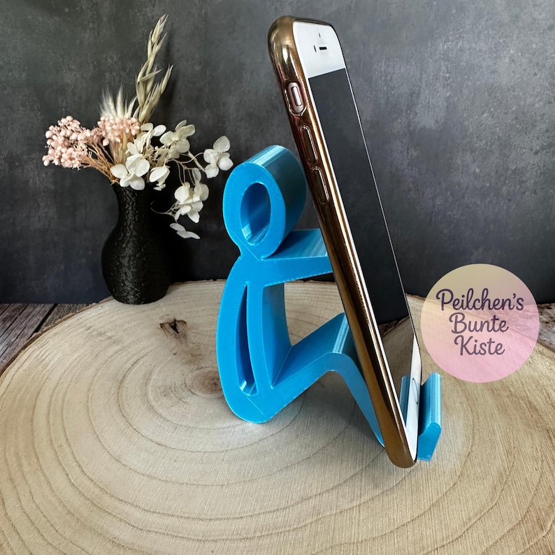 Pu&ograve; includere: Un supporto per telefono blu brillante a forma di persona che tiene in mano un telefono dorato e nero. Il supporto &egrave; su una superficie di legno. Un vaso nero con fiori &egrave; sullo sfondo.