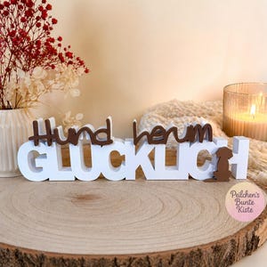 Könnte beinhalten: Ein weiß-braunes Holzschild mit dem deutschen Schriftzug "Hund herum GLUCKLICH" und einer kleinen braunen Hundesilhouette, die rechts am Wort GLUCKLICH befestigt ist. Das Schild besteht aus geschichteten Buchstaben in einer Blockschrift.