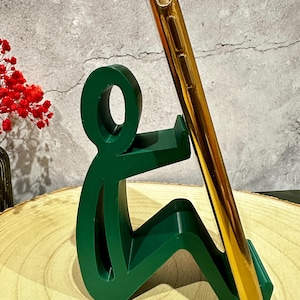 Pu&ograve; includere: Un supporto per telefono verde a forma di persona seduta, che tiene un telefono dorato. Il supporto &egrave; su una superficie di legno, con uno sfondo grigio. Fiori rossi sono visibili sullo sfondo.
