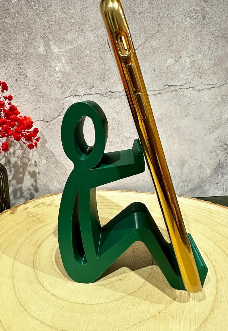 Pu&ograve; includere: Un supporto per telefono verde a forma di persona seduta, che tiene un telefono dorato. Il supporto &egrave; su una superficie di legno, con uno sfondo grigio. Fiori rossi sono visibili sullo sfondo.