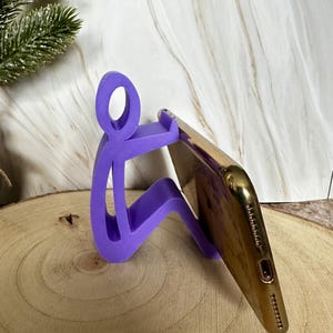 Pu&ograve; includere: Un supporto per telefono viola a forma di persona che tiene in mano uno smartphone dorato. Il supporto &egrave; realizzato in un materiale solido ed &egrave; progettato per tenere un telefono ad angolo. Il supporto &egrave; su una superficie di legno.