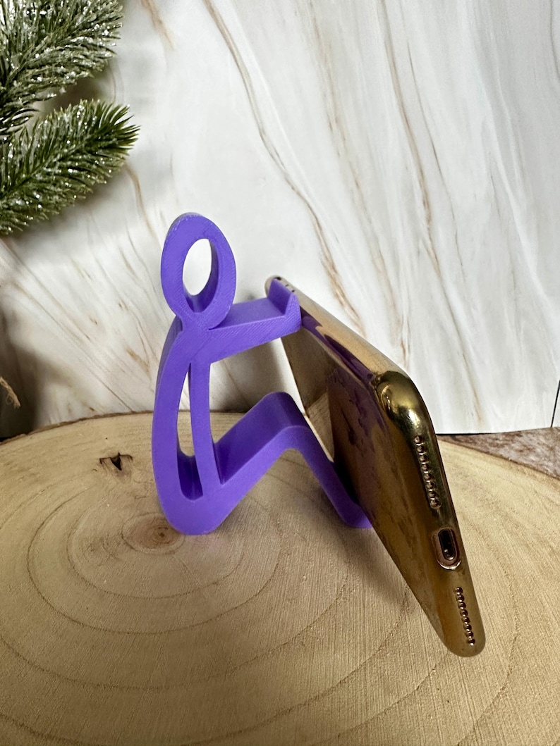 Pu&ograve; includere: Un supporto per telefono viola a forma di persona che tiene in mano uno smartphone dorato. Il supporto &egrave; realizzato in un materiale solido ed &egrave; progettato per tenere un telefono ad angolo. Il supporto &egrave; su una superficie di legno.