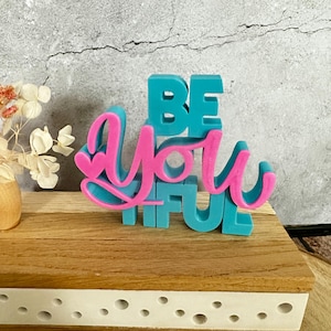 Könnte beinhalten: Ein teal- und rosa-farbenes Holzschild mit der Aufschrift "BE YOU TIFUL". Die Buchstaben "BE" und "FUL" sind teal, während "YOU" rosa ist. Das Schild steht auf einer Holzoberfläche mit weißem Rand.