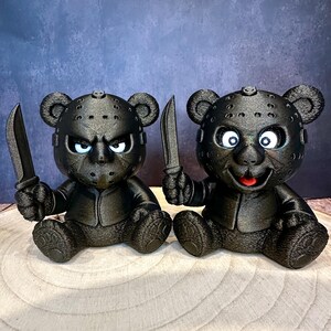 Könnte beinhalten: Zwei schwarze Bärenfiguren, die jeweils ein Messer halten. Die Bären haben eine strukturierte Oberfläche und tragen Hockeymasken. Ein Bär hat einen wütenden Ausdruck, während der andere ein lächelndes Gesicht hat. Die Figuren sitzen auf einer Holzoberfläche.