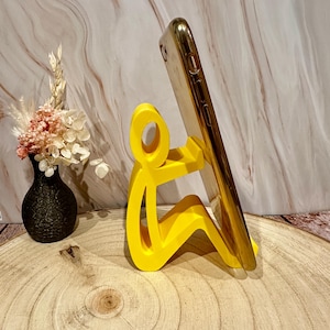 Pu&ograve; includere: Un supporto per telefono giallo brillante a forma di persona che tiene un telefono dorato. Il supporto &egrave; su una superficie di legno, con un vaso nero di fiori sullo sfondo.