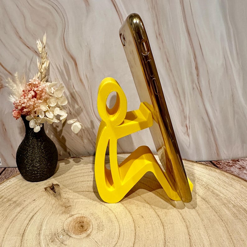Pu&ograve; includere: Un supporto per telefono giallo brillante a forma di persona che tiene un telefono dorato. Il supporto &egrave; su una superficie di legno, con un vaso nero di fiori sullo sfondo.