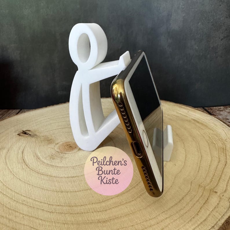 Pu&ograve; includere: Supporto per telefono bianco a forma di persona che tiene un telefono con cornice dorata e schermo nero. Il supporto &egrave; su una superficie di legno. Il testo "Peilchen's Bunte Kiste" &egrave; visibile. Supporto per cellulare.