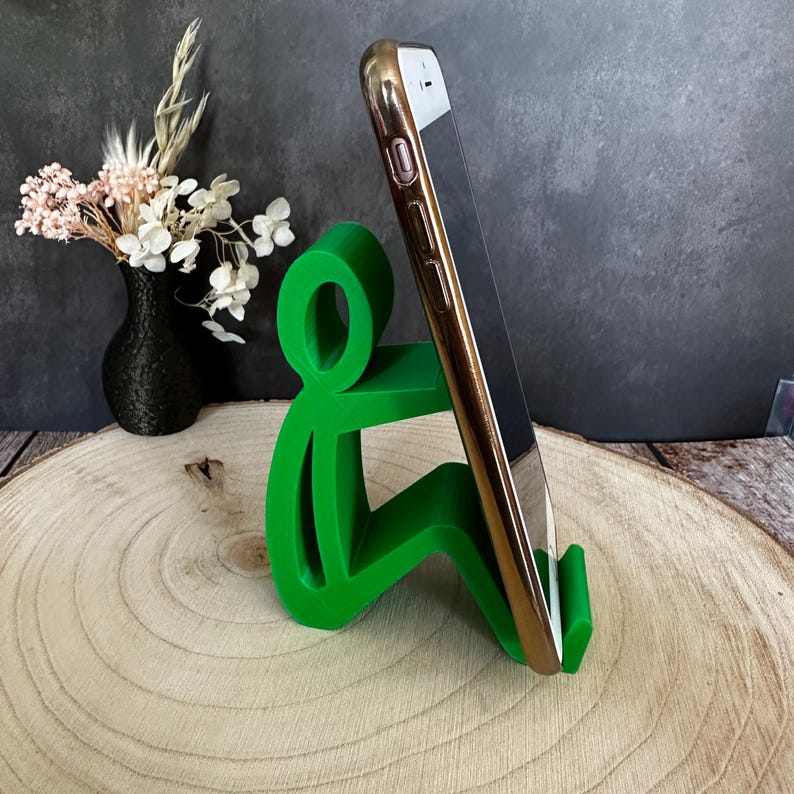 Pu&ograve; includere: Un supporto per telefono verde brillante a forma di persona seduta, che tiene in mano uno smartphone dorato. Il supporto &egrave; realizzato in un materiale solido ed &egrave; progettato per tenere un telefono in posizione verticale. Lo sfondo include un vaso nero con fiori.