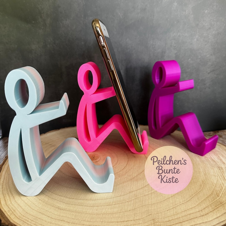 Pu&ograve; includere: Tre supporti per telefono colorati a forma di figure sedute. Uno &egrave; azzurro, uno &egrave; rosa che tiene un telefono d'oro e il terzo &egrave; viola. I supporti sono su una superficie di legno con uno sfondo scuro. Il supporto rosa tiene uno smartphone dorato.