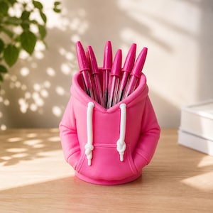 Könnte beinhalten: Ein leuchtend rosa Stifthalter in Form eines Hoodies, mit weißen Kordeln und einer Vordertasche. Der Halter ist mit rosa Stiften gefüllt. Der Stifthalter steht auf einer Holzoberfläche.