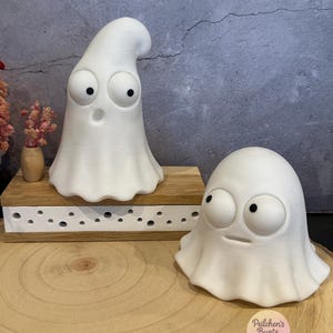 Könnte beinhalten: Zwei weiße Geisterfiguren mit großen, runden Augen und schwarzen Pupillen. Ein Geist ist groß und aufrecht mit offenem Mund, während der andere kürzer und rundlicher mit einem geraden Mund ist. Ein kleines rosa Blumenarrangement in einer Vase.