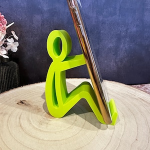 Pu&ograve; includere: Un supporto per telefono verde brillante a forma di persona seduta, che tiene in mano uno smartphone dorato. Il supporto &egrave; realizzato in un materiale solido e si trova su una superficie di legno. Lo sfondo &egrave; blu scuro.